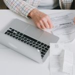 Mãos organizam documentos sobre uma mesa diante de um laptop.