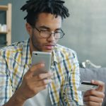 Homem usa um smartphone e um cartão de crédito enquanto faz uma compra online em casa.