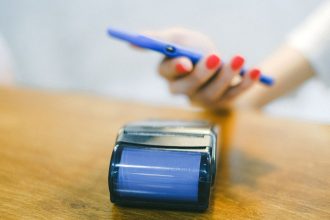 Mão aproxima um smartphone de um terminal de pagamento para realizar uma transação sem contato.
